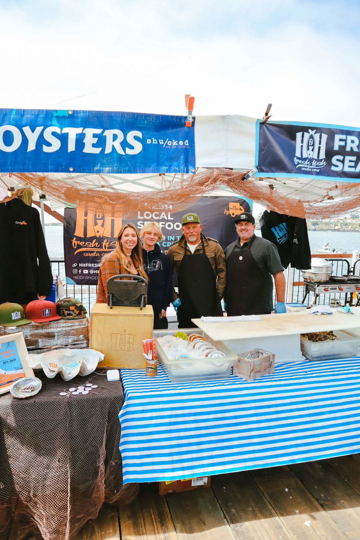 Heidi, Seb, Hans, and DJ (left to right) at a local seafood market stall, showcasing fresh oysters and seafood products under a blue 'OYSTERS' banner with branded merchandise displayed on a table with a striped blue tablecloth.