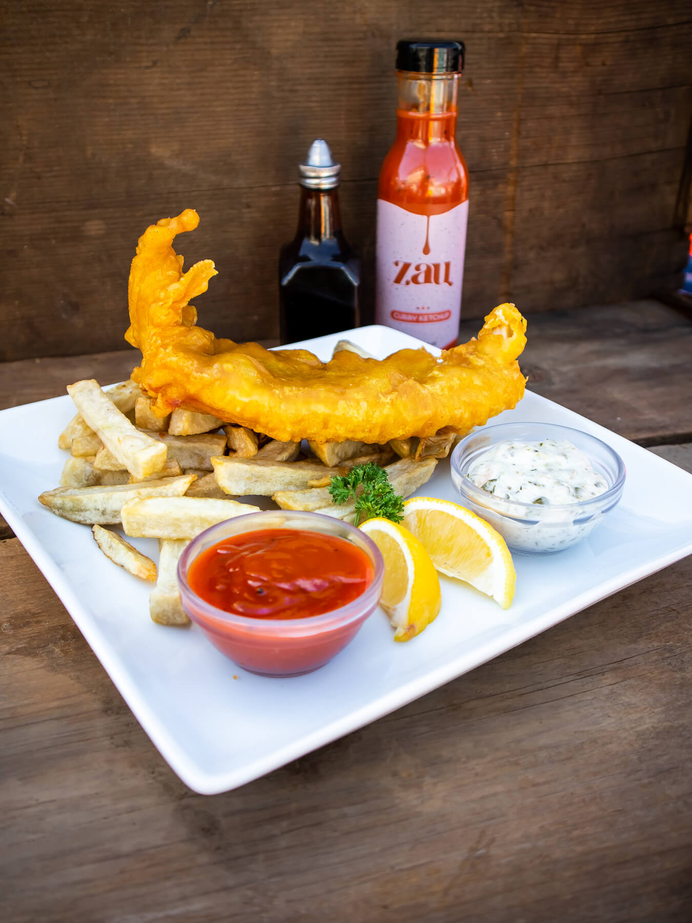 Plated fish and chips. Fish is sitting on top of chips. Two sauces are in front (first: Zau second: Tartar sauce) and bottle of Zau and bottle of Vinegar is in the background