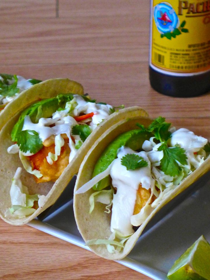 Close up of two fish tacos with white sauce, cilantro, avocado, and lime